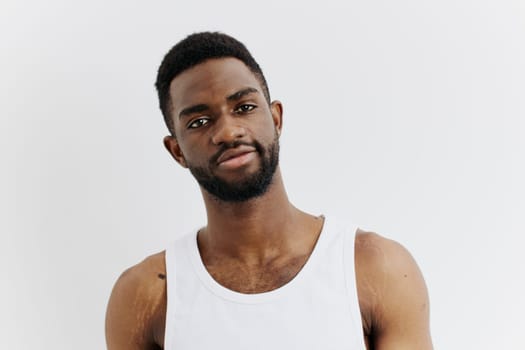 A confident young man in a white tank top stands with arms crossed against a plain studio background, exuding self-assurance.