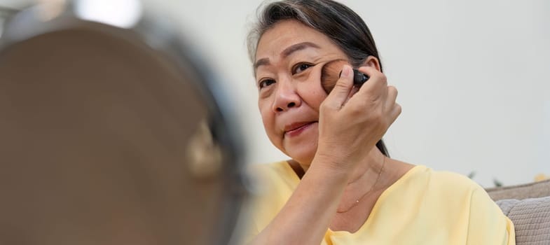 An older woman applies foundation in front of a mirror, showcasing the beauty of age diversity and self-expression through makeup.