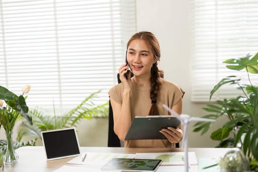 A young woman engaged in a phone call, discussing eco-friendly initiatives while surrounded by plants and sustainability documents in her workspace.