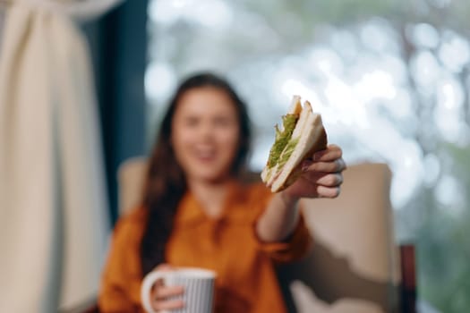 A young woman with long hair smiles as she holds a tasty sandwich and a coffee mug, set in a cozy indoor space with warm tones