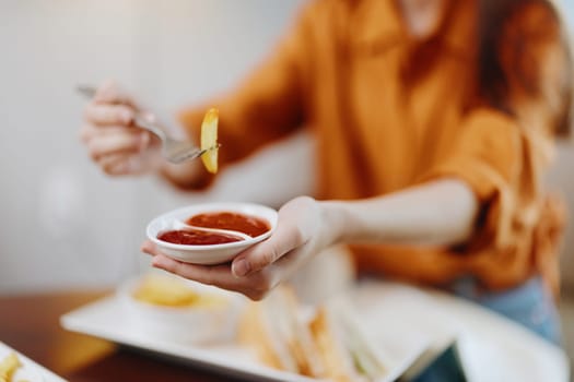 A smiling woman in an orange shirt holds a plate of fries and dips them in ketchup, reflecting joy and casual dining in a warm, modern environment