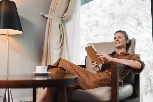 A relaxed young woman, dressed in a stylish brown jumpsuit, smiles while reading a book in a modern living room with a warm color palette