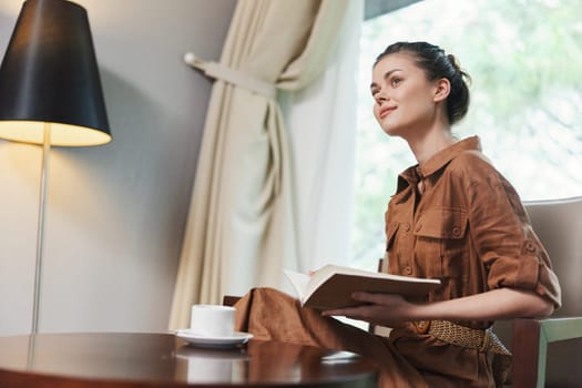 A young woman in a brown dress sits quietly with a book, immersed in reading by the window with soft, natural light creating a cozy and stylish atmosphere