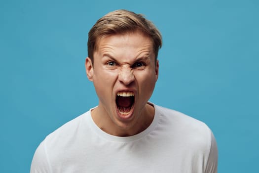 Portrait of frustrated young man with short brown hair, expressing anger or rage through shouting with open mouth, wearing casual white t-shirt, against plain blue background. People emotion concept