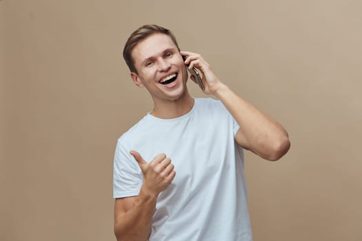 Cheerful smiling man engaging in a phone call on mobile device with a thumbs-up gesture, casual attire, light brown hair, in a neutral beige background. Modern lifestyle and communication concept