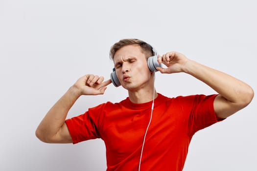 Photographer captures a relaxed young man enjoying music with over-ear headphones, wearing a bright red t-shirt, making a playful facial expression, standing against a white background. Concept: enjoyment of music and leisure time.