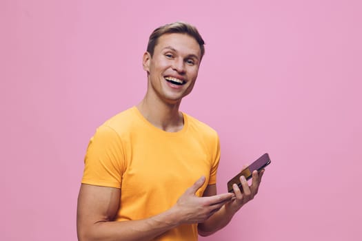Cheerful young man holding smartphone and smiling, dressed casually in yellow t-shirt, standing against pink background. Happy, technology, communication, casual style, male, young adult, lifestyle concept