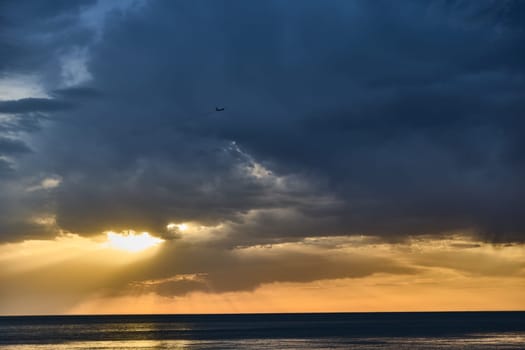dramatic sky over calm sea with sunset and airplane in twilight evening scene showing dark clouds and golden sunlight rays piercing through the clouds creating a moody atmospheric view at the ocean horizon