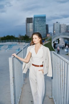 business woman in white outfit wearing blazer standing outdoors by metal fence with modern cityscape background, conveying confidence and professionalism