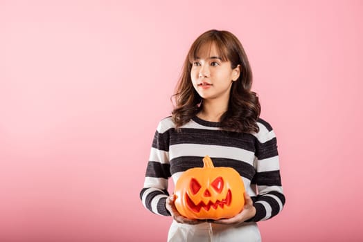 Halloween-themed studio shot, an Asian woman holds a variety of model pumpkins, including a ghost-shaped one, on a pink background. Her cheerful costume sets the tone for a fun and spooky Halloween.