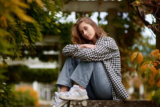 A young woman with wavy brown hair and blue eyes poses in a serene autumn park, sitting on a rustic stone wall surrounded by lush green and orange foliage. She wears a black and white checkered shirt over a white top, paired with light blue jeans and white sneakers. Her confident and relaxed expression, combined with soft natural lighting, creates a harmonious and stylish outdoor portrait. This image is ideal for autumn fashion promotions, lifestyle blogs, seasonal advertising, and social media campaigns.