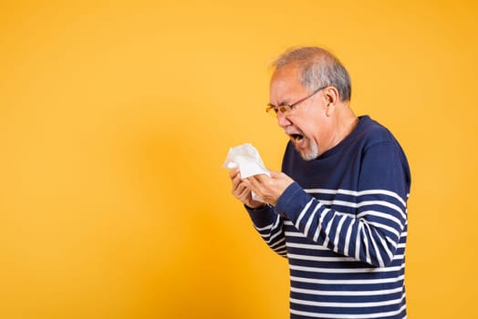 Portrait senior old man blowing nose with tissue studio shot isolated on yellow background, Asian elder man cold having flu and sneezing from sickness virus problem use tissue, pensioner unwell