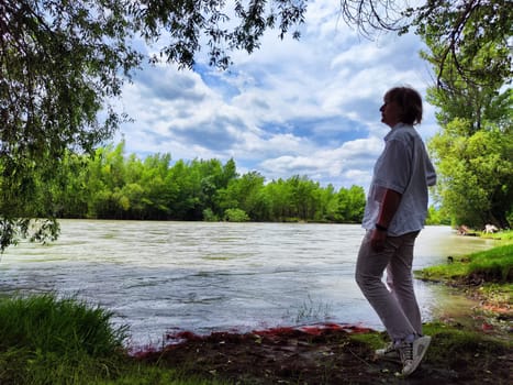 A middle-aged blonde woman stands by a quiet river, appreciating nature on a serene day with soft clouds above.
