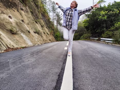 Blonde woman expresses happiness with arms outstretched while standing on a quiet road surrounded by greenery.