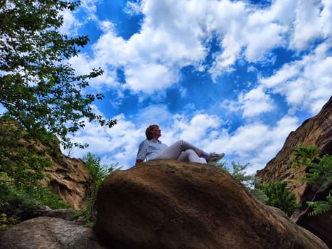 Blonde woman in casual attire sits peacefully on a large rock, appreciating the beautiful clouds and lush greenery around her.