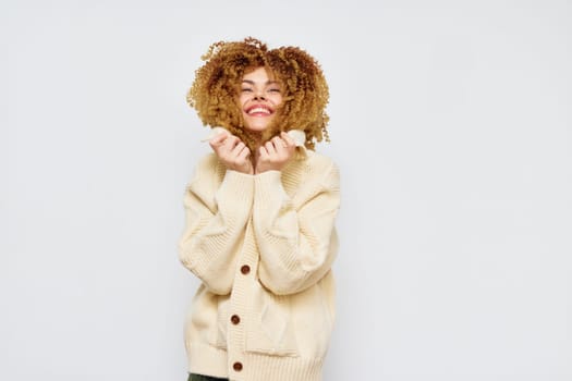 A cheerful young woman with curly hair and a warm sweater smiling widely while holding her hair, creating a playful and light hearted atmosphere The clean white background enhances her joyful expres