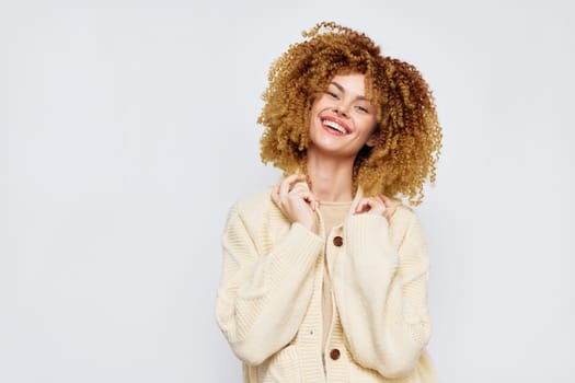 A joyful woman with curly blonde hair wearing a cozy light colored cardigan smiles warmly against a subtle gray background Ideal for portraying happiness, warmth, and fashion