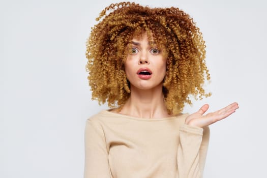 A surprised young woman with voluminous curly blonde hair looks astonished, standing against a soft light background Her expression showcases a mix of shock and curiosity, inviting viewer engagement
