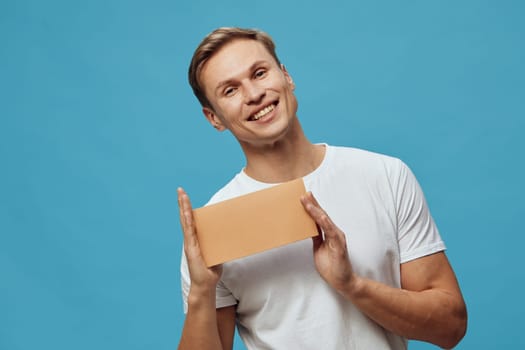Cheerful young man with short hair wearing a white T-shirt, smiling and holding a blank cardboard sign, against a solid blue background, casual pose for lifestyle or marketing visuals. Concept: positivity and communication.
