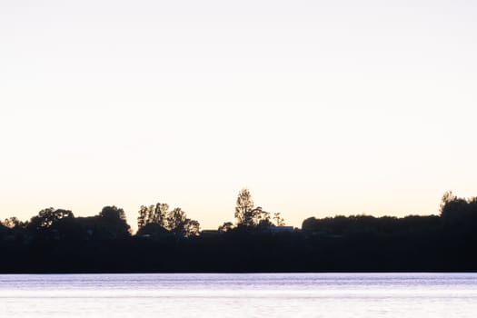 Land and trees in silhouette on horizon at sunrise across bay.