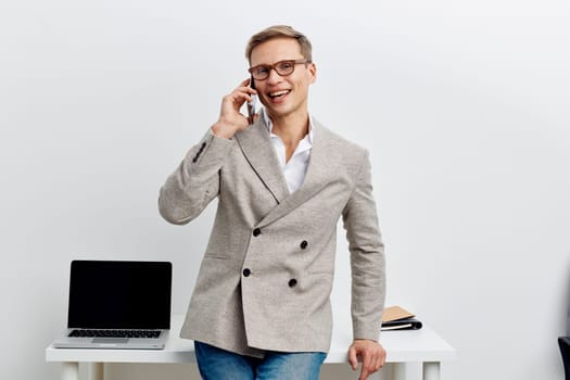 smiling man talking phone wear casual business jacket and glasses standing near laptop on desk in bright room modern workspace remote work digital nomad concept communication connection