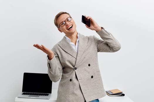Cheerful woman wearing glasses and a blazer, talking on her smartphone with a big smile, standing in a bright office space with minimalistic desk, laptop, and notebooks, conveying confidence, communication, technology, professional environment. Concept: positive work attitude.