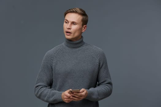 Young man with short brown hair in gray turtleneck sweater holding smartphone, posing indoors with neutral gray background. Casual, modern look conveying communication, technology, and everyday lifestyle concept.