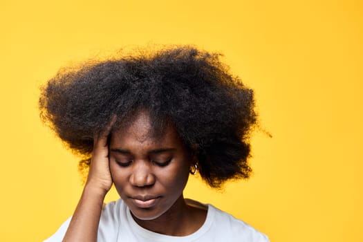 An African woman with voluminous curly hair shows signs of stress and worry The bright yellow backdrop enhances the emotional tone, emphasizing her feelings