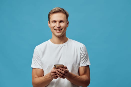 Confident young man with a bright smile holding a mobile phone, standing against plain blue backdrop. Casual outfit and positive vibe highlight modern technology usage, upbeat atmosphere, lifestyle, communication, happiness, connection, studio photography.