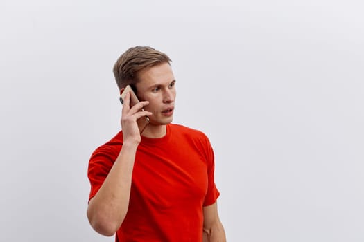 Young caucasian man talking on mobile phone wearing red t-shirt with serious expression isolated on white background. Modern communication and digital nomad lifestyle concept with mobile technology used for global connection.
