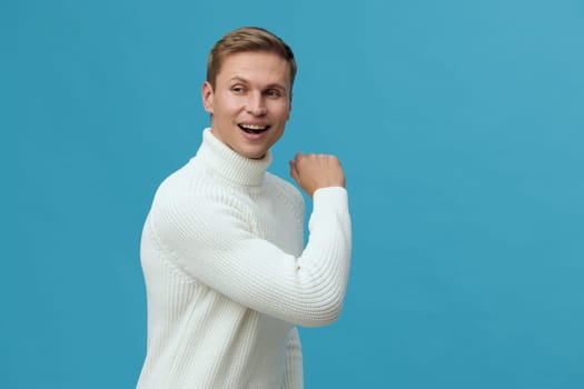Smiling man in white sweater looking back over shoulder on plain blue background. Young caucasian man expressing joy and confidence in casual wear. Concept: digital nomad lifestyle with remote work and technology.
