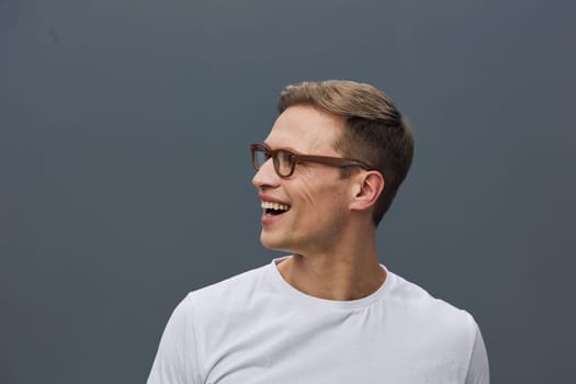 Happy man with short brown hair and glasses wearing a white casual t-shirt, smiling and looking to the side, with a plain gray studio background. Concept: joyful, casual, positive mood.