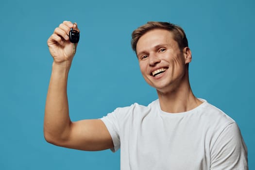 Happy young man with short brown hair and casual white t-shirt presenting car key in hand on blue background. Studio portrait capturing excited emotion, celebration, confidence, achievement, lifestyle, success, joy concept.