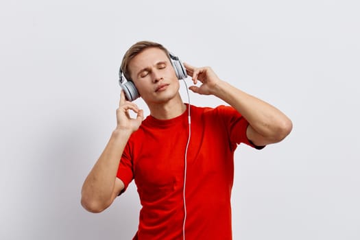 Young caucasian man in red t-shirt wearing headphones with closed eyes enjoy music and relax with mental health and gadgets concept. Positive lifestyle and digital detox idea with white background studio portrait.