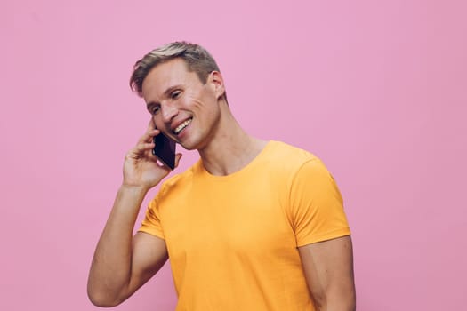 Happy smiling man engaging in a phone conversation on a mobile device, dressed in a vibrant yellow t-shirt, standing in front of a pink backdrop. Modern casual style, youthful energy, positive mood, lifestyle concept, studio setting, colorful scheme, relaxation