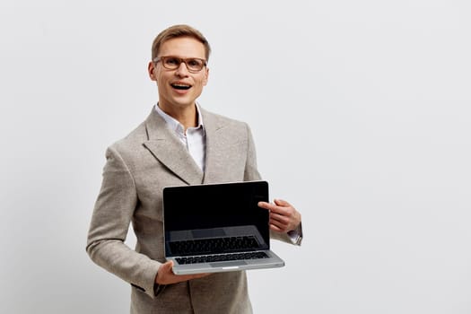 Cheerful man with glasses dressed in a gray blazer holding a laptop computer, standing confidently against a plain white backdrop. Suitable for technology, business, remote work, professional, modern workplace, success, workplace concept