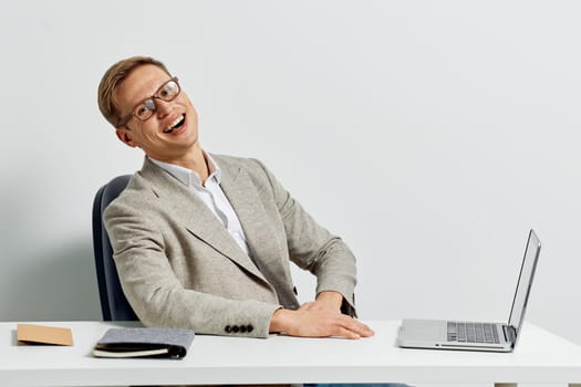 Smiling man working with laptop wearing casual jacket and eyeglasses sitting at white desk against light background. The cheerful young man represents a digital nomad engaged in remote work and modern technology use. Concept: digital nomad lifestyle