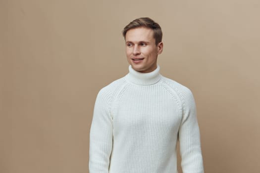 Friendly man dressed in white turtleneck sweater smiling and looking to the side against a plain beige background in a studio shot. Warm cozy clothing, casual style, cheerful mood concept