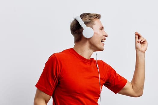 Cheerful young man in casual red clothing wearing white headphones, expressing happiness while dancing or listening to music, excitation and fun, positive energy, plain white background, studio shot, lifestyle theme