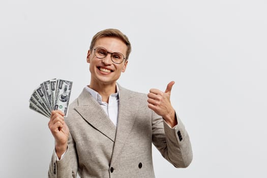Happy man dressed in professional beige suit with glasses, smiling and displaying cash in hand, pointing thumbs up, isolated on white background, emphasizing wealth, success, prosperity, finance, celebration concept.