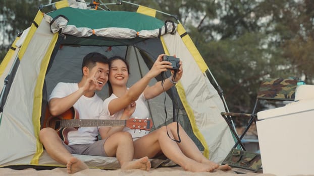 Young loving couple pretty while camp taking photo by camera while sitting at the camping tent on beach in holiday vacation time at sunset, Young Asian group woman and man in summer travel outdoor