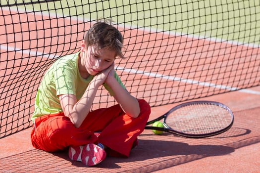 The boy lost the tennis competition. The child sits at the net with a racket. Psychology of sports in childhood.