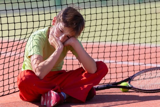 The boy lost the tennis competition. The child sits at the net with a racket. Psychology of sports in childhood.
