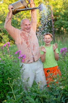 A grandfather waters flowers with a watering can while his grandson watches and laughs. The scene is peaceful and serene, father and son enjoying the simple pleasure of tending to their garden.