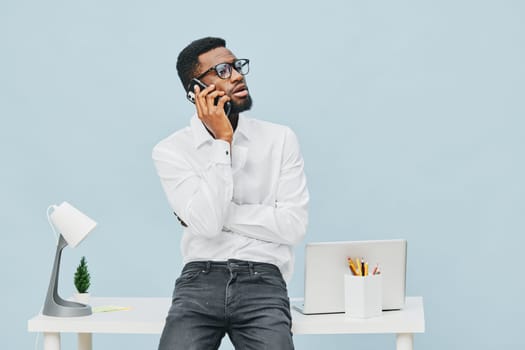 Relaxed young African American man in casual white shirt and glasses engaging in a phone call at contemporary office workspace with laptop, desk accessories, and soft blue background, demonstrating communication and productivity