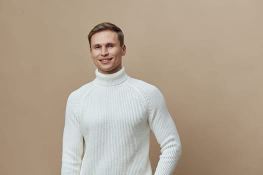 Portrait of cheerful young man with short hair, wearing cozy white knitted turtleneck sweater standing against a simple beige backdrop. The image captures a warm, friendly atmosphere perfect for fashion, lifestyle, or happiness themes. Concept: positive attitude.