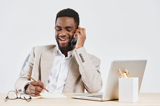 Cheerful man engaged in a phone call at his workspace in a well-lit, contemporary office environment, with a laptop, notebook, and pen in front of him. Captures professionalism, communication, and productivity in workplace setting. Concept: office communication and modern work