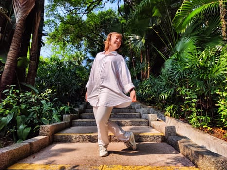 A middle-aged woman with blonde hair playfully poses on garden steps, surrounded by lush greenery, enjoying a sunny day outdoors.