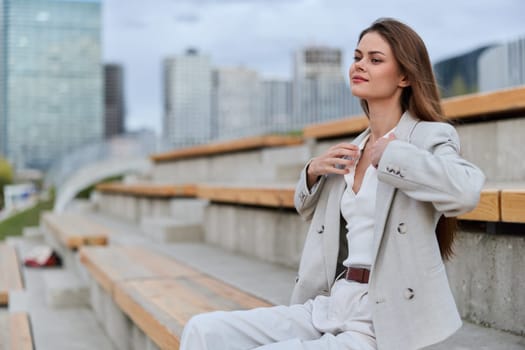 Confident woman in beige business suit sits relaxed on urban benches with modern cityscape behind, showing calm and professional attitude in spring business setting