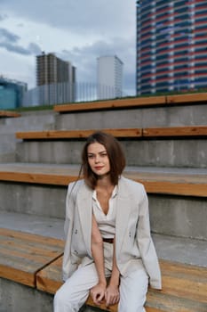 A poised businesswoman in a tailored suit is seated on wooden steps, exuding confidence and elegance, with contemporary buildings in the background, ideal for corporate themes.